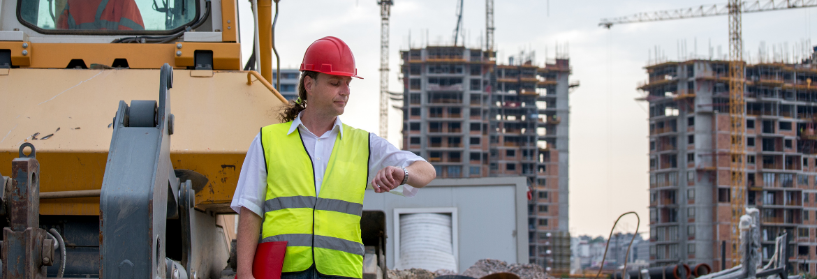 Construction site engineer stands next to a bulldozer in front of the construction site, looking at the time on his watch