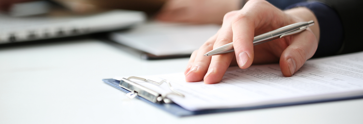 Hand of businessman in suit filling and signing with silver pen partnership agreement form clipped to pad closeup.