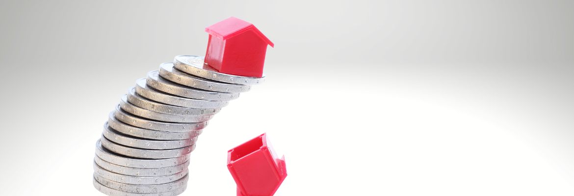 A red house on a slanting stack of coins. Below one house that already fell.